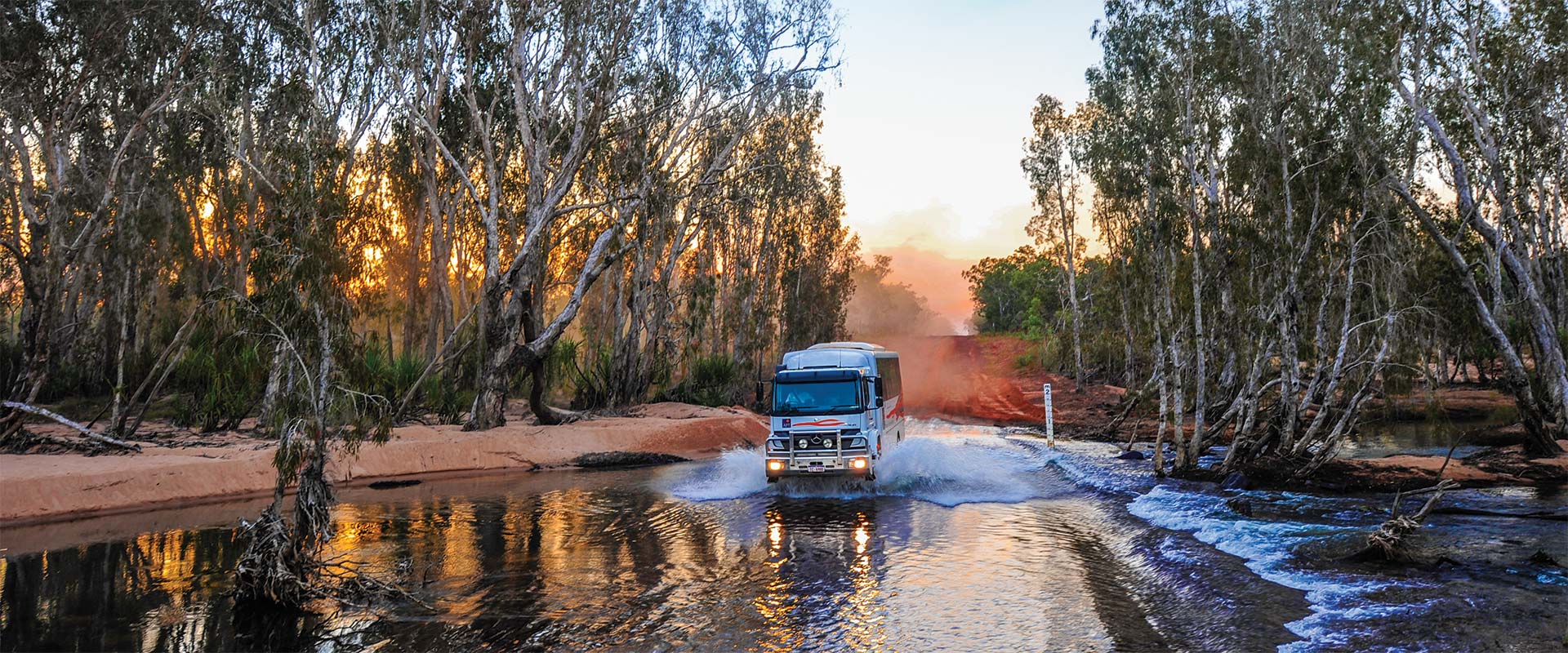 4WD vehicle crossing river surrounded by red earth and gum trees, WA